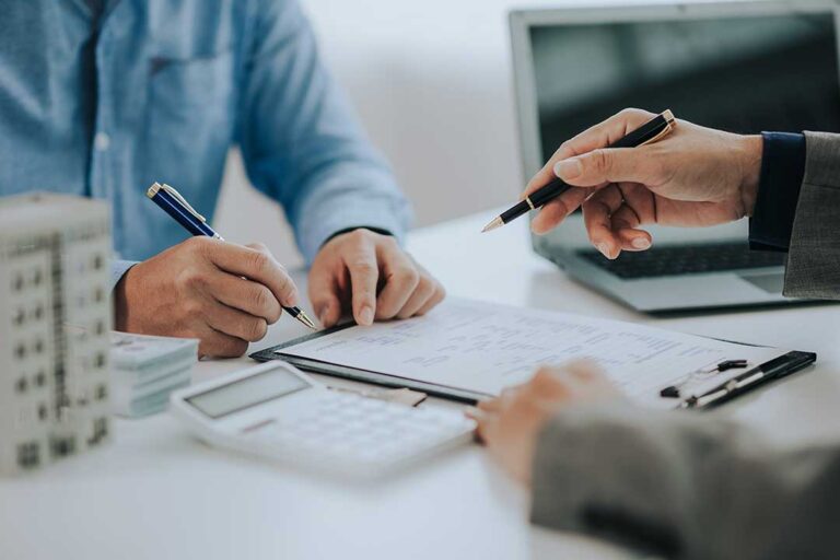 A person reviews a home insurance policy at a kitchen table with property paperwork nearby.